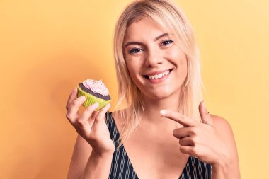 Sarışın, güzel bir kadın izole edilmiş sarı arka planda tatlı çikolatalı kek yerken mutlu mesut elini ve parmağını göstererek gülümsüyor.