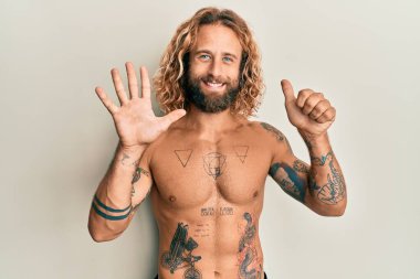 Sakallı, uzun saçlı, gömleksiz, altı numaralı parmaklarını gösteren dövmeleri olan yakışıklı bir adam. Kendinden emin ve mutlu bir şekilde gülümsüyor.. 