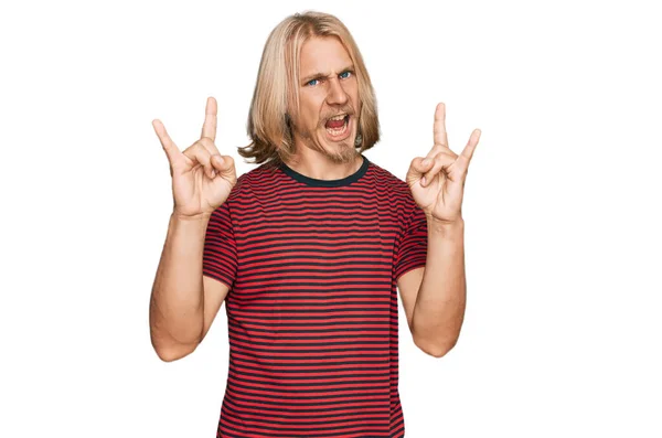 金髪の長髪の白人男性が手を上げて岩のシンボルをやって狂気の表情で叫んでカジュアルな縞模様のTシャツを着ています 音楽スター 重い概念は — ストック写真