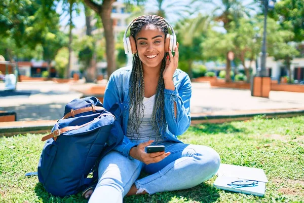 若いアフリカ系アメリカ人の学生の女性が笑顔で大学のキャンパスで草の上に座って音楽を聞いて幸せ — ストック写真