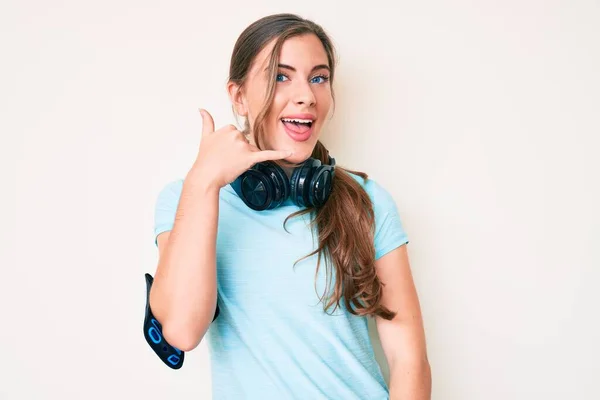 漂亮的年轻的高加索女人穿着运动服 用耳机笑着用手和手指做电话手势 就像在电话里说话一样 交流概念 — 图库照片