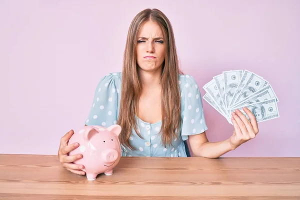 Young Blonde Girl Holding Dollars Piggy Bank Sitting Table Skeptic — Stock Photo, Image