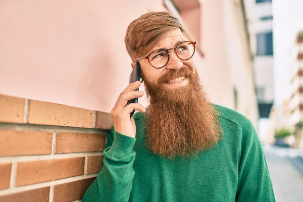 Kızıl saçlı genç İrlandalı adam mutlu bir şekilde gülümsüyor ve şehirdeki akıllı telefondan konuşuyor..