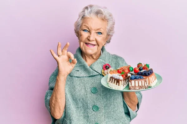 ケーキのスライスでプレートを保持シニア白髪の女性は指でOkサインを行います 笑顔優しいジェスチャー優れたシンボル — ストック写真
