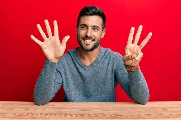 自信と幸せな笑顔ながら テーブルの上に座って 指の数8を示すと指摘カジュアルなスタイルを身に着けているハンサムなヒスパニック系の男 — ストック写真