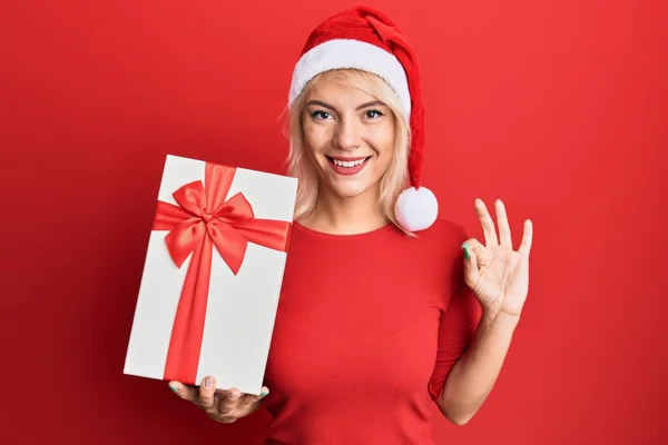 Young Blonde Girl Wearing Christmas Hat Holding Gift Doing Sign — Stock Photo, Image