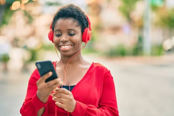 Young african american woman smiling happy using smartphone and headphones at the city.