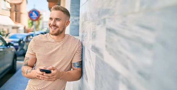 Genç İrlandalı mutlu bir şekilde gülümsüyor. Şehrin duvarına yaslanmış akıllı telefon kullanıyor..