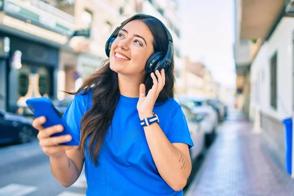 Genç İspanyol kadın şehirde akıllı telefon ve kulaklık kullanarak mutlu bir şekilde gülümsüyor..