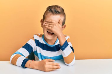 Gündelik giysiler giymiş sevimli beyaz bir çocuk masaya oturmuş gülümsüyor ve elleri yüzlerinde şaşkınlıktan gözleri kapatıyordu. kör kavram. 