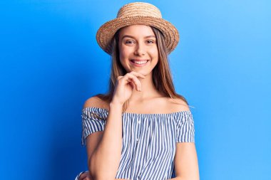 Yazlık şapka ve çizgili tişört giyen genç ve güzel bir kız kameraya kollarını çapraz yapıp çenesine dokunarak gülümsüyor. Olumlu düşünmek. 