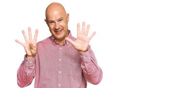 Orta yaşlı, kel bir adam, günlük kıyafetler giyip dokuz numaralı parmaklarıyla işaret ederken, kendinden emin ve mutlu bir şekilde gülümsüyor.. 