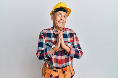 Tamirci üniforması giyen İspanyol kıdemli bir adam. Elleriyle yalvarıyor ve dua ediyor. Yüzünde umut ifadesi var. Çok duygusal ve endişeli. Yalvarıyor. 