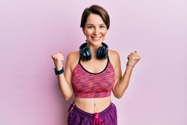 Kısa saçlı, spor giyimli ve kulaklıklı genç esmer kadın kolları açık ve gözleri açık bir şekilde başarıyı kutluyor ve şaşırıyor. Kazanan konsept. 