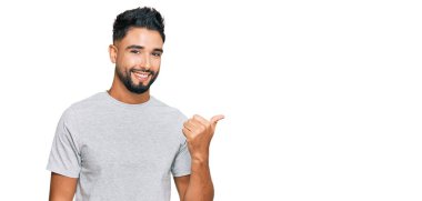 Sakallı, günlük gri tişörtlü, mutlu yüzüyle gülümseyen ve baş parmağıyla yüzünü gösteren genç bir adam.. 