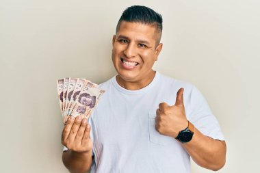 500 Meksika pezosu tutan genç Latin adam mutlu ve olumlu gülümsüyor, başparmağıyla mükemmel ve onay işareti yapıyor. 