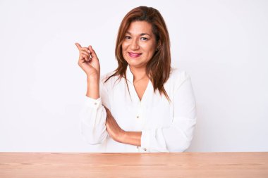 Orta yaşlı esmer, Latin iş kadını. Masada oturan, yüzünde kocaman bir gülümseme olan, eli ve parmağı kameraya bakan gündelik beyaz gömlekli kadın.. 