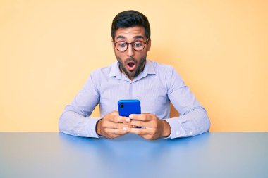 Genç İspanyol adam masada oturmuş akıllı telefon kullanıyor. Korkmuş ve şaşkın bir şekilde ağzı açık bekliyor. 