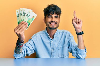 Genç İspanyol adam İsrail şekellerini elinde tutarak masaya oturmuş bir fikir ya da soruyla gülümsüyor ve mutlu bir yüzle işaret ediyor. 