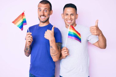 Genç eşcinsel çift ellerinde gökkuşağı lgbtq bayrakları, gülümseyen mutlu ve pozitif, başparmak yukarı mükemmel ve onay işareti ile 