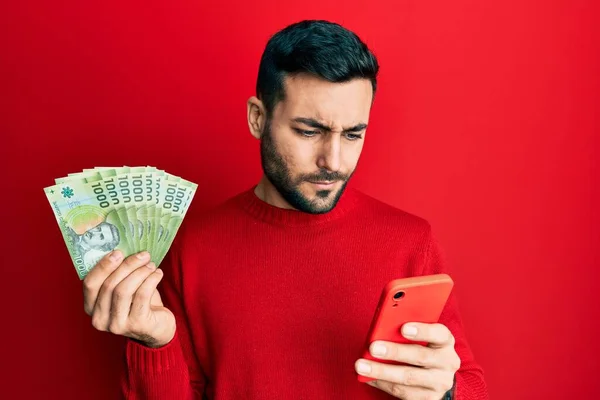 Young Hispanic Man Using Smartphone Holding Chilean Pesos Banknotes Skeptic — Stock Photo, Image