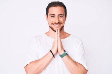 Gündelik beyaz tişört giyen genç yakışıklı ellerini birleştirip af diliyor kendinden emin bir şekilde gülümsüyor.. 