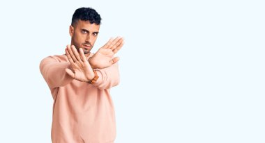 Gündelik kıyafetler giyen genç İspanyol adam. Reddedilmiş yüz ifadesi. Kollar ve avuçlar çapraz. 