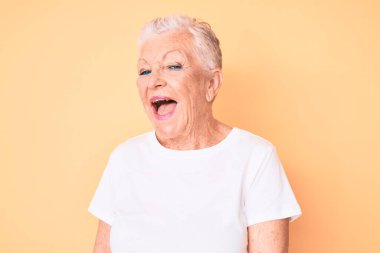 Senior beautiful woman with blue eyes and grey hair wearing classic white tshirt over yellow background winking looking at the camera with sexy expression, cheerful and happy face. 