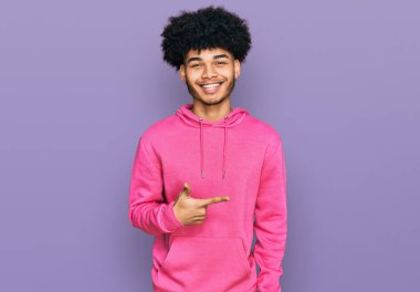 Afro saçlı, neşeli pembe kazaklı, yüzünde gülümsemesi olan, mutlu ve doğal bir ifadeyle elini ve parmağını kaldırmış Afro Amerikalı genç bir adam. 