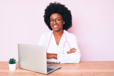 Doktor steteskobu giyen genç Afro-Amerikalı kadın bilgisayarla çalışıyor. Kameraya bakarken kollarını kavuşturup gülümsüyor. pozitif kişi. 