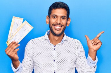 Elinde biniş kartı tutan genç latin adam gülümsüyor mutlu bir şekilde elini ve parmağını yana doğru gösteriyor. 