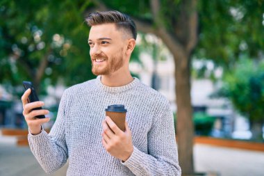 Genç beyaz adam akıllı telefon kullanıyor. Parkta kahve içiyor..