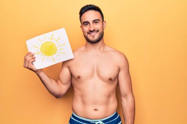 Tatilde sakallı, mayo giyen, elinde güneş alan bir kağıt tutan yakışıklı genç adam. Pozitif ve mutlu görünüyor. Kendine güvenen bir gülümsemeyle dişlerini gösteriyor.
