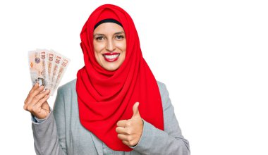 İslami tesettürlü güzel bir kadın elinde 5 kilo banknotla mutlu ve pozitif gülümsüyor, başparmağıyla mükemmel ve onay işareti yapıyor. 