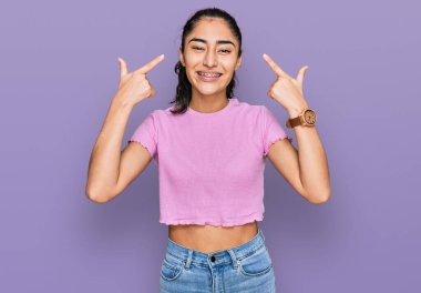 Diş teli takan, sıradan kıyafetler giyen, iki eliyle başını gösteren İspanyol bir genç kız. Harika bir fikir ya da düşünce, iyi bir hafıza. 