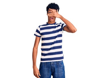 Genç, Afro-Amerikan bir adam günlük giysiler giymiş. Şoka girmiş, yüzünü ve gözlerini kapamış, utanmış bir ifadeyle parmaklarının arasından bakıyor.. 