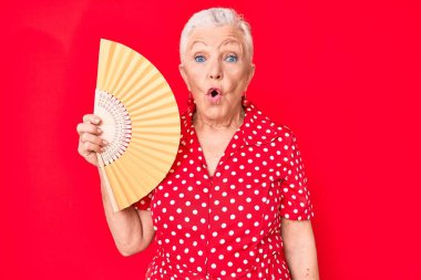 Senior beautiful woman with blue eyes and grey hair waving hand fan cooling air in summer scared and amazed with open mouth for surprise, disbelief face 