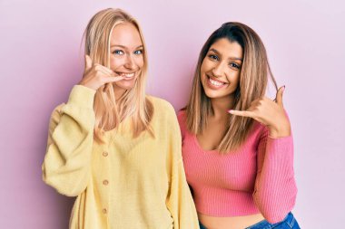 Pembe arka planda yan yana duran iki arkadaş gülümseyerek el ve parmaklarıyla telefonla konuşuyormuş gibi davranıyorlar. iletişim kavramları. 