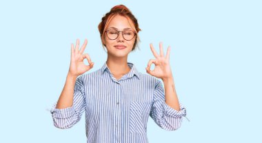 Genç kızıl saçlı kadın günlük kıyafetler ve gözlük takıyor rahatlıyor ve gözleri kapalı gülümsüyor parmaklarıyla meditasyon hareketi yapıyor. yoga konsepti. 