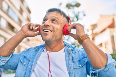 Genç beyaz adam şehirde kulaklık takarak mutlu bir şekilde gülümsüyor..