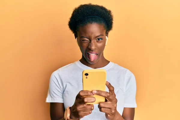 年轻的非洲裔美国女孩用智能手机和耳机伸出舌头 带着滑稽的表情高兴 — 图库照片