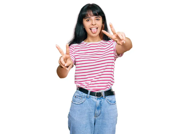 両手の指を見せて舌で微笑むカジュアルな服を着た若いヒスパニック系の女の子が勝利サインをしています — ストック写真