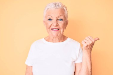 Son sınıf güzeli, mavi gözlü, gri saçlı, sarı arka planda klasik beyaz tişört giyen, gülümseyen, gülümseyen ve baş parmağıyla yüzünü gösteren bir kadın.. 