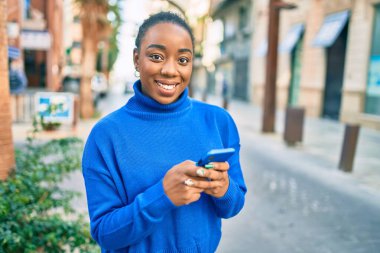 Genç Afrikalı Amerikalı kadın şehirde akıllı telefon kullanarak mutlu bir şekilde gülümsüyor..