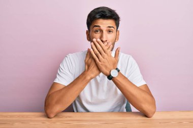 Masada oturan sıradan beyaz tişört giyen genç yakışıklı adam yanlışlıkla elleri ile ağzını kapatırken şok oldu. gizli kavram. 