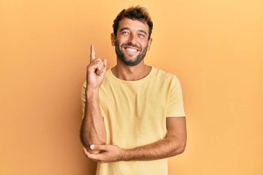 Sarı arka planda sıradan sarı tişörtler giyen sakallı yakışıklı bir adam. Yüzünde kocaman bir gülümseme, eli ve parmağı kameraya bakıyor.. 