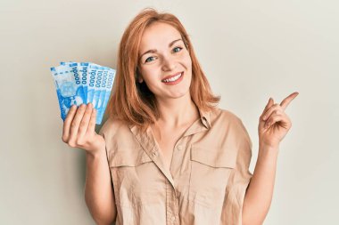 On bin Şili pezosu tutan beyaz bir kadın mutlu bir şekilde elini ve parmağını yana uzatarak gülümsüyor. 