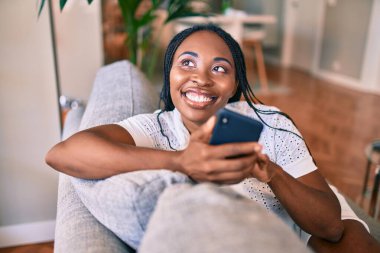 Genç Afrikalı Amerikalı kadın gülümseyerek evde akıllı telefon kullanarak mutlu mesut oturuyor.