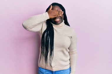 Saçları örgülü, sıradan bir süveter giyen, gülümseyen ve yüzünde şaşkınlıktan gözleri kapayan genç siyahi bir kadın. kör kavram. 