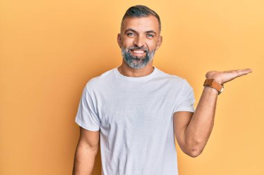 Orta yaşlı, beyaz tişört giyen, neşeli gülümseyen, avuç içi kameraya bakan yakışıklı bir adam.. 